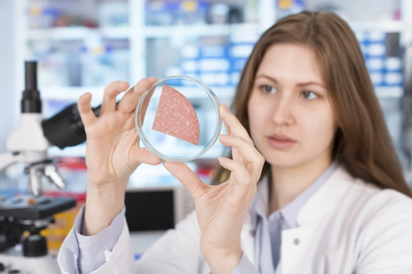 Kobieta w białym fartuchu laboratoryjnym trzyma szalkę Petriego z fragmentem mięsa laboratoryjnego, w tle widoczny mikroskop oraz rozmyte laboratorium.