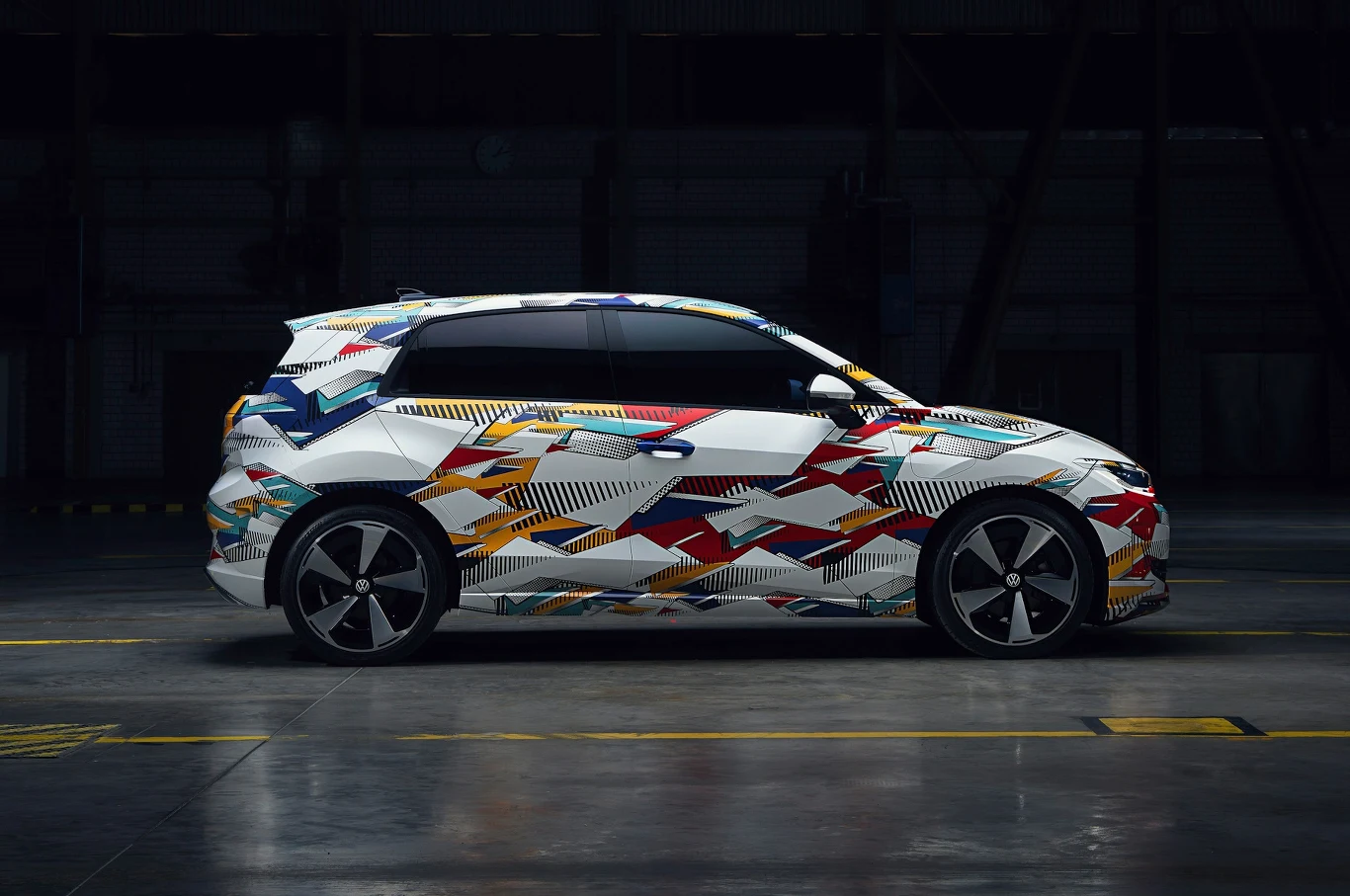 Samochód osobowy z nowoczesnym i kolorowym wzorem kamuflażu na karoserii, zaparkowany w dużej, słabo oświetlonej hali przemysłowej.