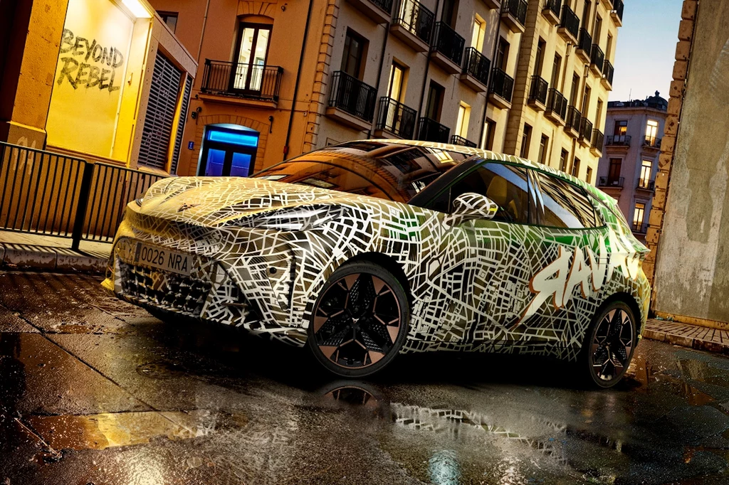 Cupra Raval Samochód osobowy z dynamicznym wzorem graficznym na karoserii, zaparkowany na mokrej ulicy miejskiej w nocy, w tle oświetlone kamienice i neonowy napis na ścianie.