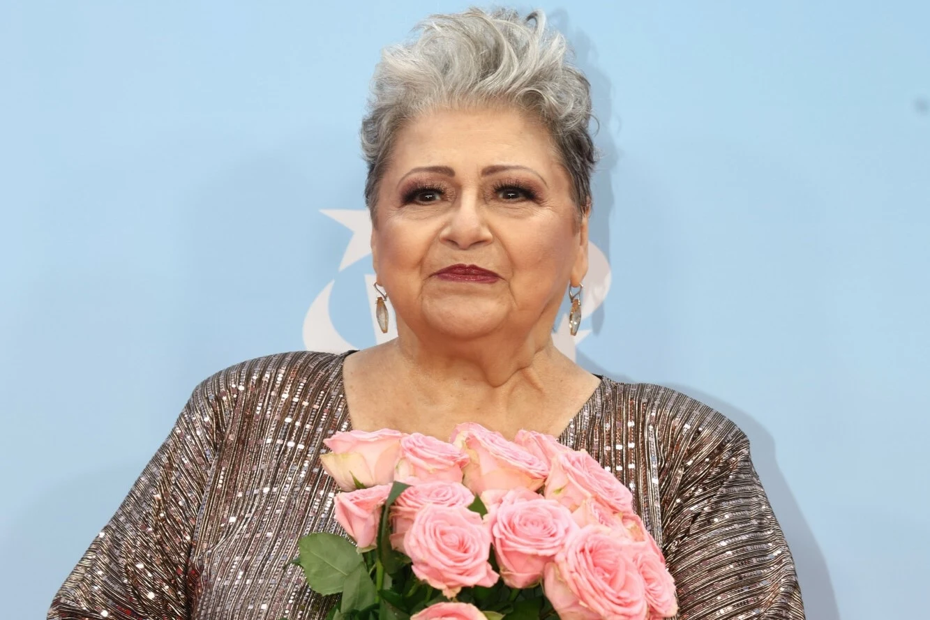 Ewa Bem przeżywa żałobę po stracie córki i męża Starsza kobieta o siwych włosach w eleganckiej brokatowej sukience trzymająca bukiet świeżych, różowych róż, w tle jasnoniebieska ścianka.