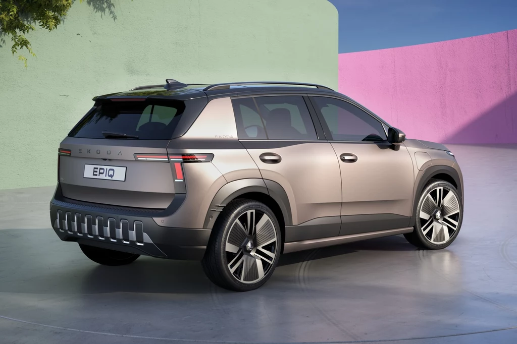 Skoda Epiq Nowoczesny, srebrny SUV marki Škoda z widocznym oznaczeniem modelu EPIQ stoi na odkrytym, jasnym placu z pastelowym, geometrycznym tłem; samochód prezentuje dynamiczne linie nadwozia, duże felgi i ciemne przeszklenia.