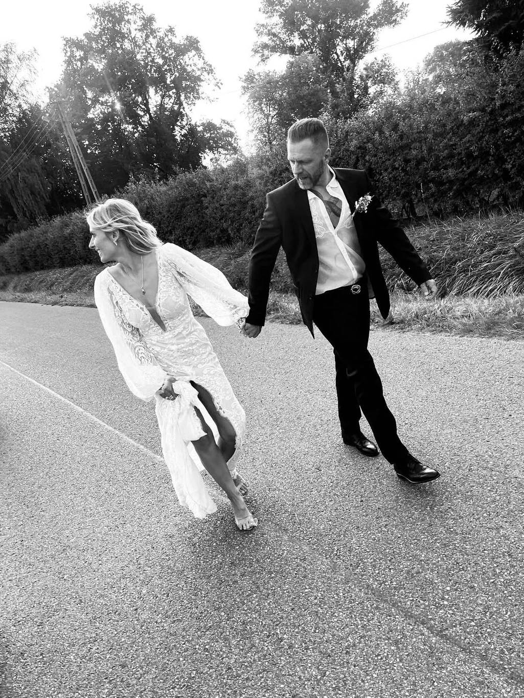 Zdjęcie z ceremonii ślubnej, które Patrycja Markowska opublikowała na swoim Instagramie Para młoda w eleganckich strojach trzyma się za ręce, idąc boso po asfaltowej drodze, panna młoda w koronkowej sukni ślubnej i pan młody w garniturze, w tle zieleń i drzewa.