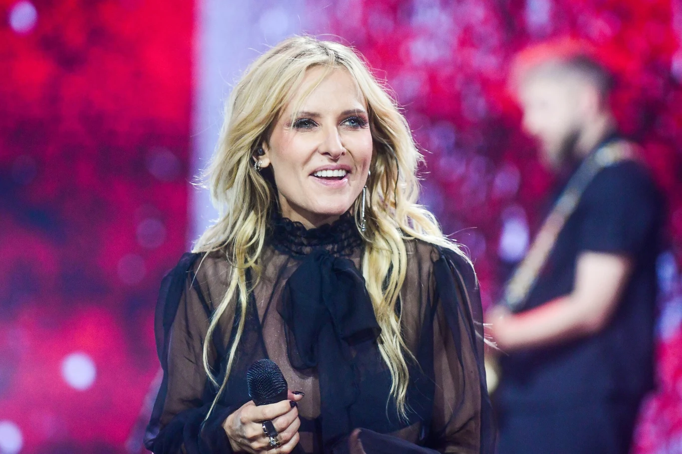 Patrycja Markowska opublikowała wzruszający post w pierwszą rocznicę ślubu Uśmiechnięta kobieta z długimi blond włosami, ubrana w czarną, przezroczystą bluzkę z kokardą, trzyma mikrofon podczas występu na scenie, w tle rozmyta sylwetka muzyka oraz czerwone światła sceniczne.