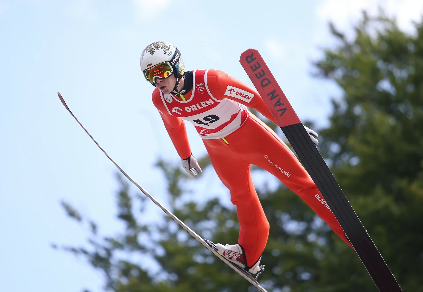 Skoczek narciarski w czerwonym kombinezonie i kasku w locie nad śnieżnym podłożem, dynamiczna pozycja na tle błękitnego nieba i zielonych drzew, widoczne profesjonalne narty z napisem VAN DEER.