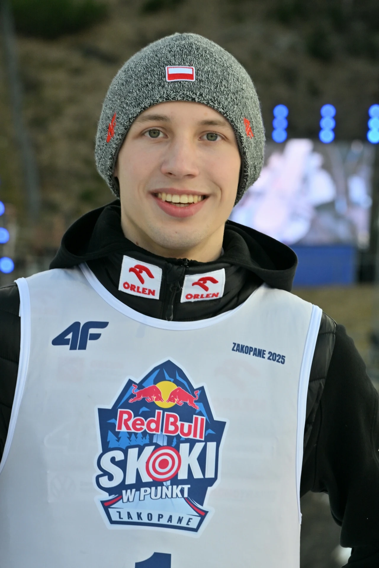 Uśmiechnięty młody mężczyzna w sportowej kamizelce z logotypem Red Bull Skoki w Punkt oraz czapce zimowej z naszywką flagi Polski, w tle nieostry fragment trybun i świateł stadionowych.