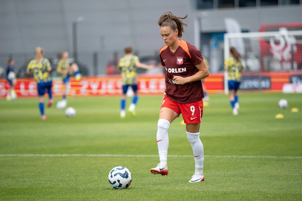 Ewa Pajor Zawodniczka w bordowej koszulce z numerem 9 na boisku piłkarskim podczas treningu lub rozgrzewki, w tle inne piłkarki oraz piłki rozłożone na murawie.