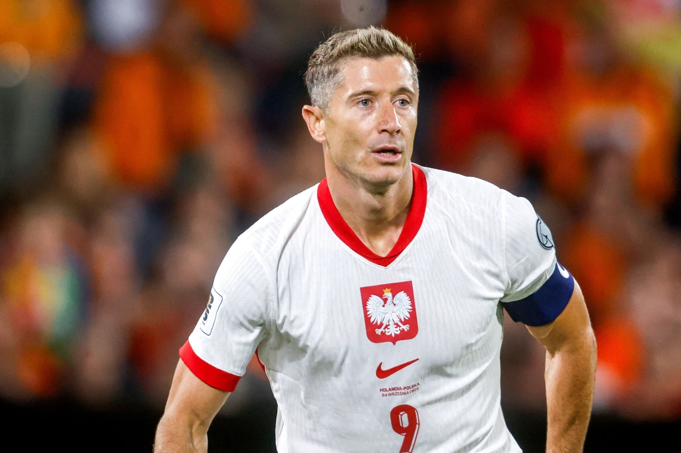 Piłkarz w białej koszulce z czerwonym orłem na piersi oraz numerem 9 w trakcie meczu, z założoną opaską kapitana, na tle zamazanych trybun z kibicami.