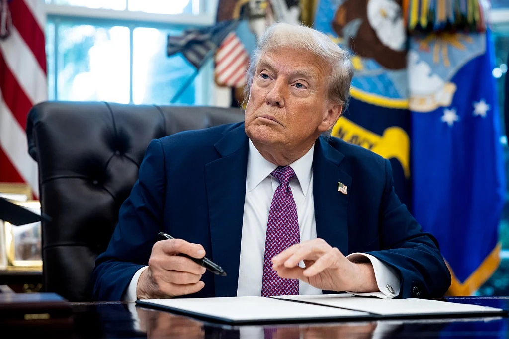 Donald Trump zapowiedział, że jest gotowy do przejścia do kolejnej fazy sankcji na Rosję Mężczyzna w garniturze siedzi przy biurku, trzyma długopis i patrzy na bok, na tle widać flagi oraz charakterystyczne elementy gabinetu.