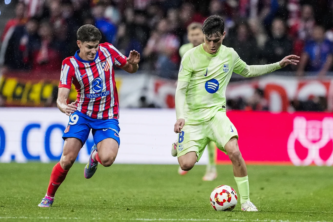 Dwóch piłkarzy walczy o piłkę podczas meczu, jeden w pasiastym czerwono-białym stroju drużyny Atlético Madryt, drugi w jasnozielonym stroju FC Barcelona, tło rozmyte z kibicami na trybunach.