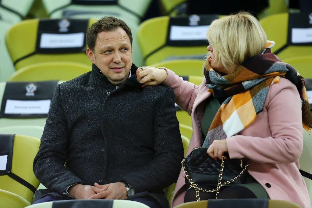 Mężczyzna w ciemnym płaszczu siedzi na stadionowych trybunach, obok niego kobieta w jasnym płaszczu i kolorowym szaliku poprawia mu kołnierz, w tle puste żółto-zielone siedzenia.