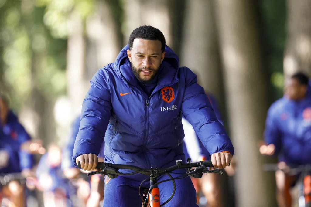 Piłkarz ubrany w niebieską kurtkę treningową z herbem reprezentacji Holandii i logiem sponsora jedzie na rowerze po alei z drzewami, w tle widać innych zawodników w podobnych strojach.