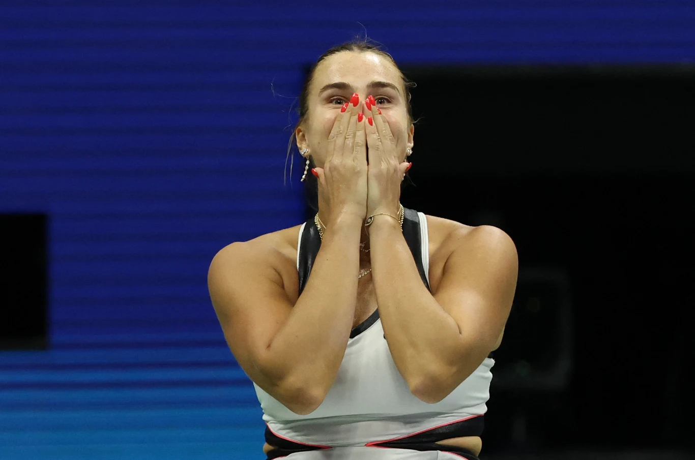 Kobieta w stroju sportowym trzyma dłonie przy ustach, okazując silne emocje, na tle niebieskiej areny sportowej.