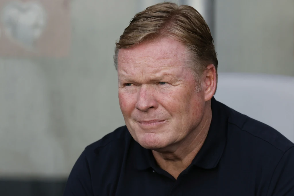 Ronald Koeman Mężczyzna w średnim wieku o jasnych włosach, ubrany w ciemną koszulkę, siedzi z poważnym wyrazem twarzy na neutralnym tle.