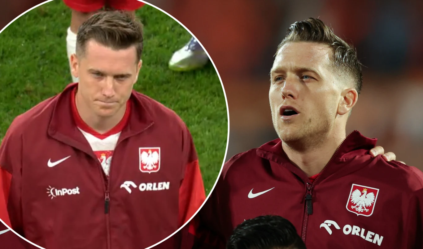 Polski piłkarz w czerwonym dresie reprezentacji Polski z logotypami sponsorów, podczas hymnu narodowego, w tle fragment murawy boiska.