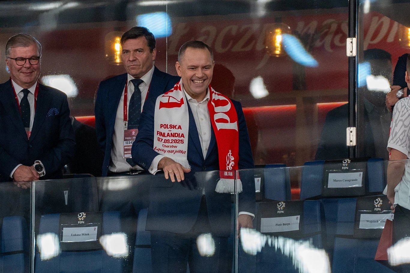 Uśmiechnięty mężczyzna w garniturze z szalikiem kibica Polski, stojący w loży honorowej stadionu, w towarzystwie innych elegancko ubranych osób, z widoczną nazwą meczu Polska – Finlandia na szaliku.