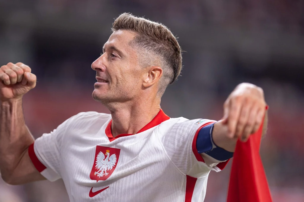 Robert Lewandowski Piłkarz w biało-czerwonej koszulce z herbem Polski, unosi rękę w geście triumfu, uśmiechnięty, w tle rozmyte trybuny stadionu.