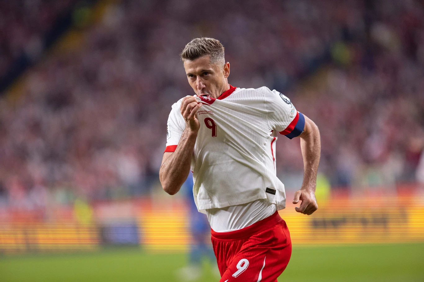 Piłkarz w biało-czerwonym stroju z numerem 9 całuje herb na koszulce, biegnąc po boisku w geście radości po zdobyciu gola, na tle pełnych trybun stadionu.