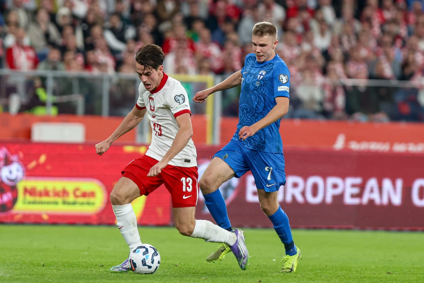 Dwóch piłkarzy rywalizuje o piłkę podczas meczu piłkarskiego na stadionie, zawodnik w biało-czerwonym stroju prowadzi piłkę, a zawodnik w niebieskim stroju próbuje go powstrzymać, w tle widoczni kibice i trybuny.