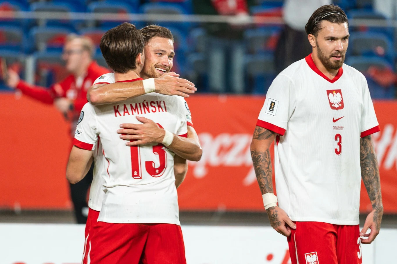 Piłkarze reprezentacji Polski w białych koszulkach świętują sukces na murawie stadionu, jeden z zawodników z numerem 13 na plecach przytula kolegę z drużyny, w tle trybuny wypełnione kibicami.