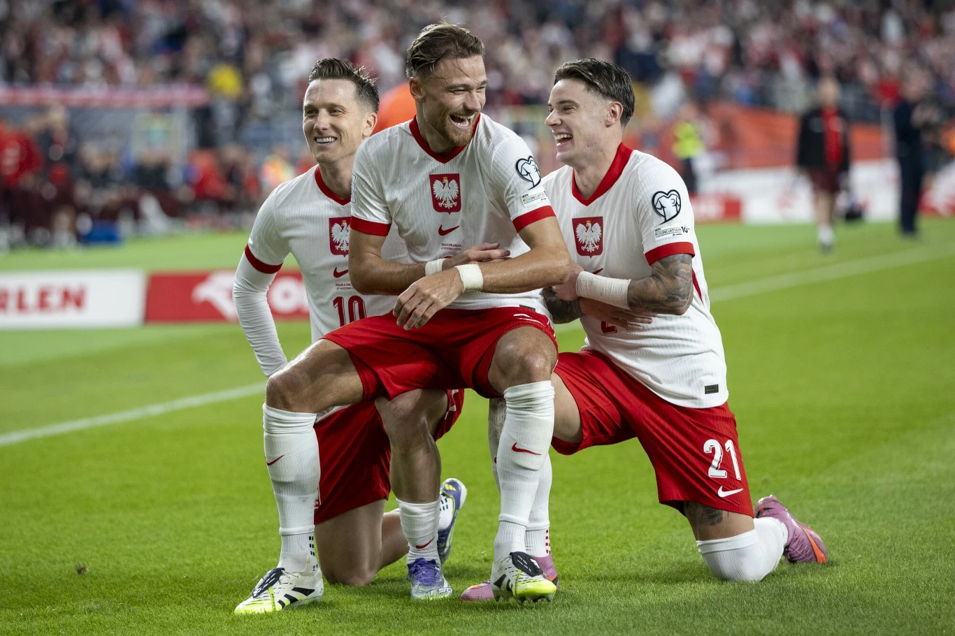 Trzech polskich piłkarzy w białych koszulkach i czerwonych spodenkach świętuje zdobycie bramki podczas meczu na zielonej murawie, wokół rozciąga się stadion pełen kibiców.