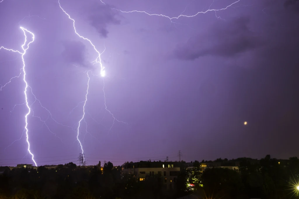 Prognoza pogody. Gdzie jest burza? W części Polski zagrzmi Jasne błyskawice rozświetlają nocne niebo nad miastem, poniżej widoczne sylwetki budynków i delikatnie oświetlone okna, a na tle nieba fragmenty ciemnych chmur.