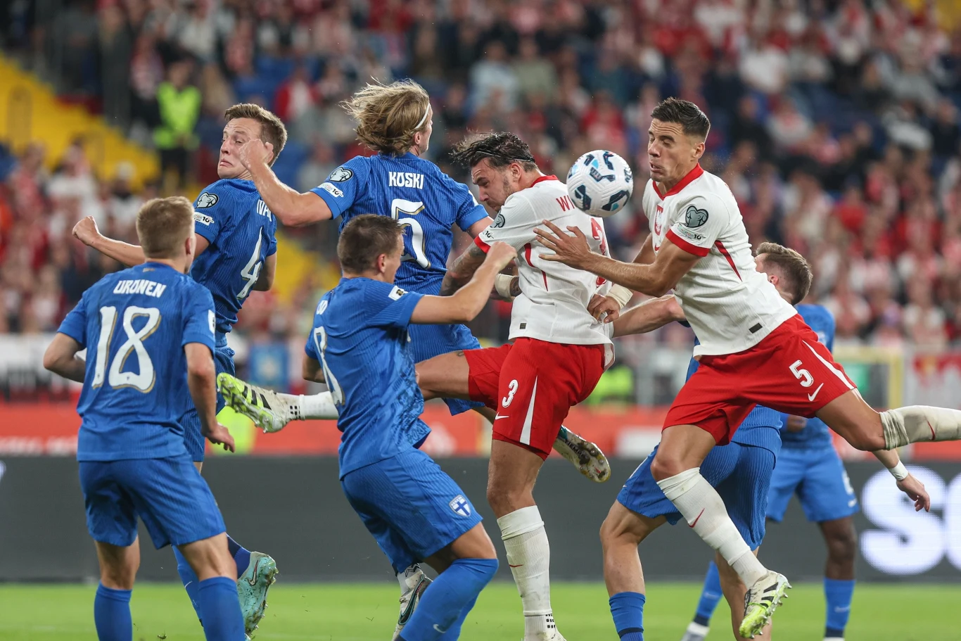 Piłkarze drużyn w niebieskich i białych strojach walczą w powietrzu o piłkę podczas meczu na stadionie, tłum kibiców w tle, intensywna sportowa rywalizacja.