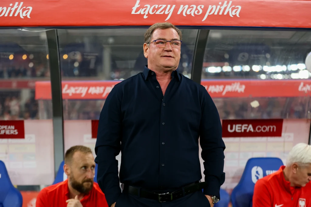 Mężczyzna w ciemnej koszuli stoi przed ławką rezerwowych na stadionie piłkarskim, za nim widoczny jest napis 'Łączy nas piłka' oraz logo UEFA, w tle dwaj mężczyźni siedzą na ławce.