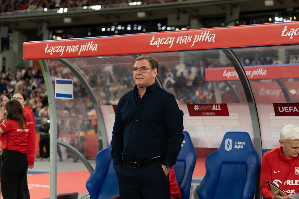 Jan Urban Trener stojący przy ławce rezerwowych na stadionie piłkarskim, wokół niego znajdują się członkowie sztabu oraz widoczna jest publiczność na trybunach.
