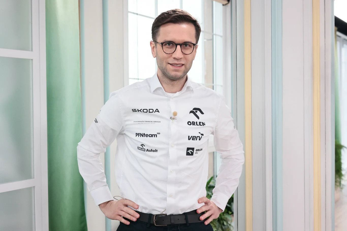 Mężczyzna w okularach ubrany w białą koszulę z licznymi logotypami sponsorów, stoi z rękami opartymi na biodrach w nowoczesnym, jasnym wnętrzu.
