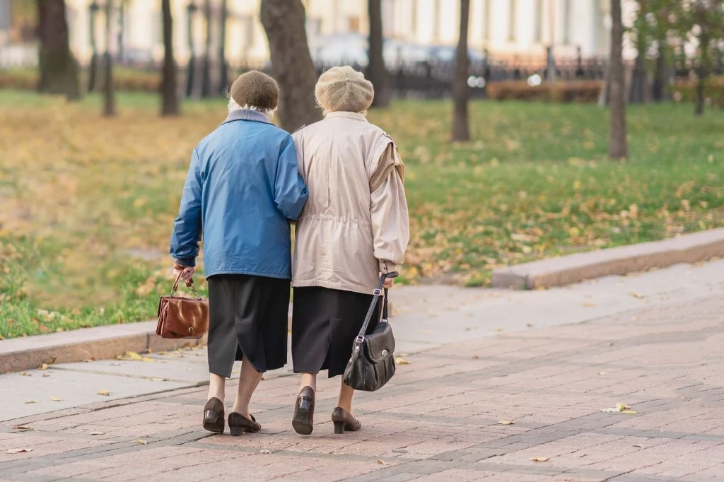 Niektórzy seniorzy otrzymają nawet o 200 zł wyższe emerytury w 2026 roku Dwie starsze kobiety idące razem chodnikiem w parku, ubrane w ciepłe płaszcze i berety, trzymające się pod rękę, każda niesie torebkę, w tle jesienne drzewa i trawniki pokryte opadłymi liśćmi.