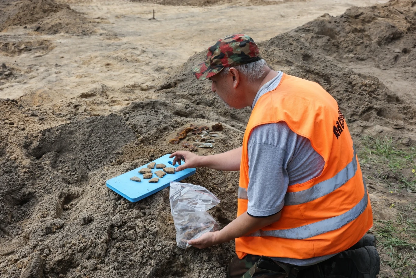 Poszukiwacze odnaleźli nie tylko fragmenty maszyny, ale też ślady z epoki brązu. Mężczyzna w kamizelce odblaskowej i czapce w militarny wzór pracuje przy wykopaliskach archeologicznych, porządkując wydobyte z ziemi artefakty na niebieskiej planszy.
