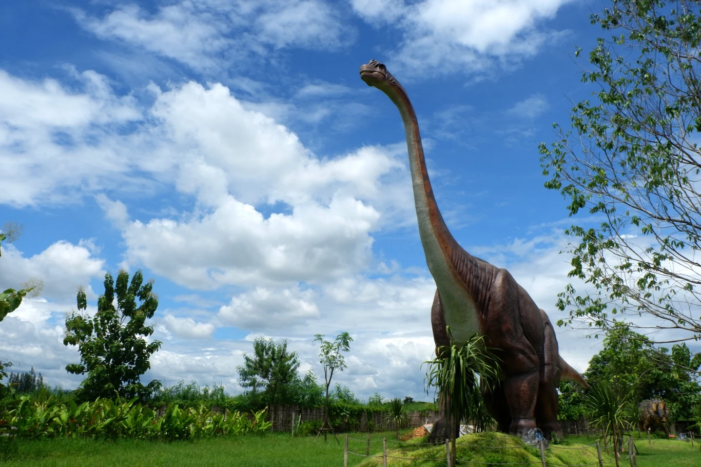 Wysoka figura dinozaura z długą szyją stoi na zielonej trawie, otoczona roślinnością i drzewami, na tle błękitnego nieba z białymi chmurami.