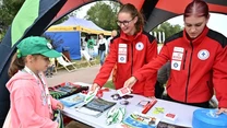 Dwie młode kobiety w czerwonych kurtkach z logo ratownictwa medycznego rozdają ulotki, broszury i gadżety na stoisku plenerowym młodej dziewczynce z zieloną czapką. Na stole widać różnorodne materiały edukacyjne i promocyjne.