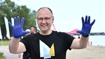 Uśmiechnięty mężczyzna w okularach, ubrany w czarną koszulkę z napisem Nieporęt i graficznym symbolem, pokazuje uniesione ręce w niebieskich, ochronnych rękawiczkach na tle nadmorskiego deptaka i plaży