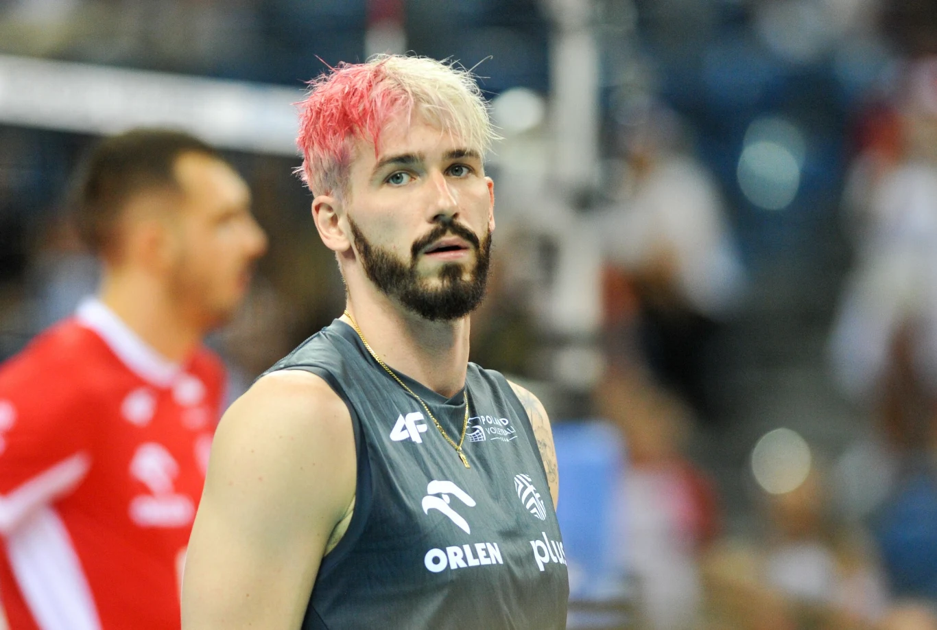 Tomasz Fornal Sportowiec z dwukolorowymi włosami ubrany w ciemny strój sportowy, w tle sylwetka innego zawodnika w czerwonym stroju siatkarskim, rozmyte trybuny i światła hali.