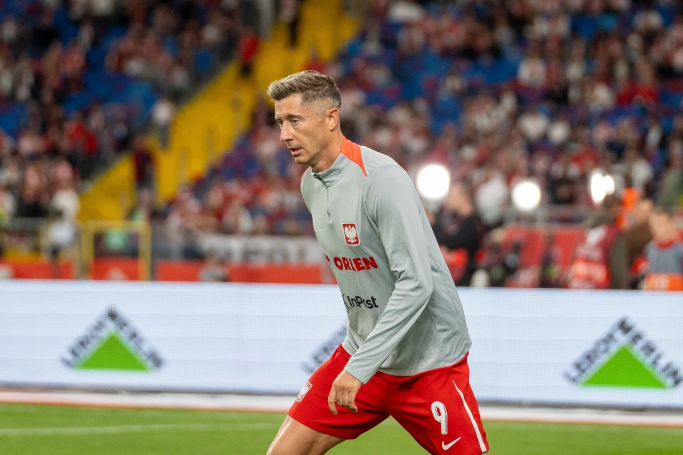Robert Lewandowski Piłkarz w szarej bluzie z numerem 9 na czerwonych spodenkach na stadionie podczas rozgrzewki, w tle rozmyta publiczność i fragment boiska.