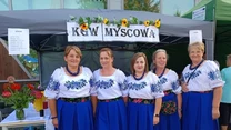 Pięć kobiet w tradycyjnych strojach ludowych stoi przed namiotem z napisem KGW Myscowa, obok stolika z kwiatami i napojami, tło stanowią dekoracje oraz inne stoisko.