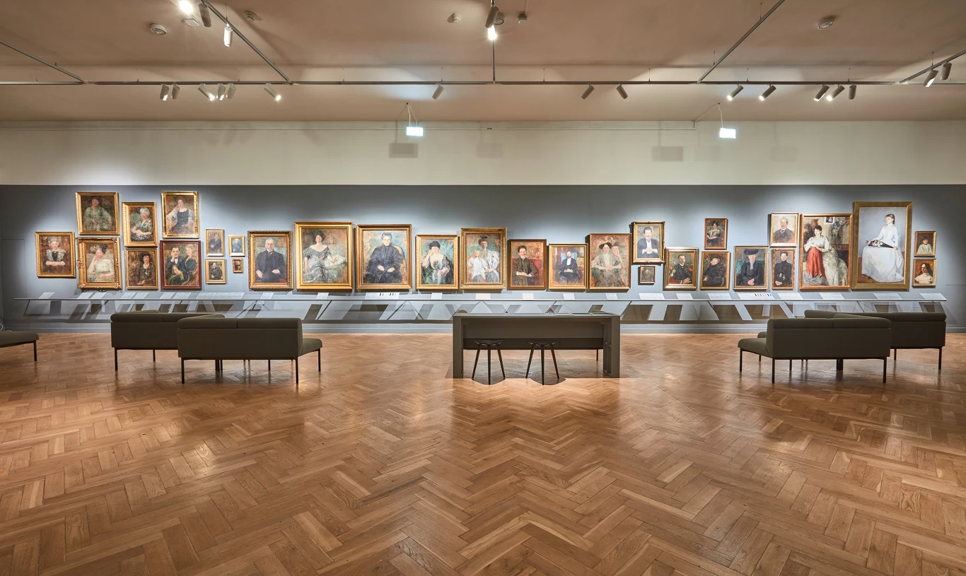 Galeria z obrazami portretowymi powieszonymi na ścianie, elegancki parkiet, ławki dla zwiedzających oraz tablice informacyjne pod obrazami, wszystko oświetlone jasnym światłem sufitowym.