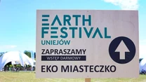 Tablica informacyjna festiwalu Earth Festival Uniejów z napisem zapraszającym do eko miasteczka na tle zielonej trawy i białych namiotów imprezowych, w słoneczny dzień.
