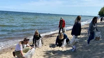 Grupa osób sprząta plastikowe i inne odpady na piaszczystej plaży nad morzem, używając worków na śmieci, wszystkim dopisuje zaangażowanie, a na horyzoncie widać spokojną wodę i niebo z lekkimi chmurami.