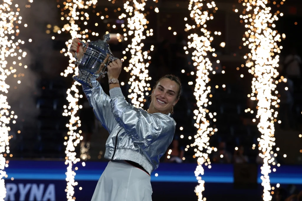 Aryna Sabalenka z trofeum US Open Tenisistka ubrana w srebrną kurtkę trzyma nad głową puchar, wokół eksplozje fajerwerków, wyrażając radość ze zwycięstwa turniejowego.