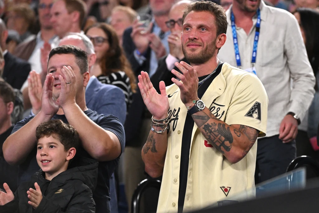 Georgios Frangulis Trzy osoby w różnym wieku stoją w tłumie i głośno klaszczą, wyrażając entuzjazm i zaangażowanie, w tle widać innych ludzi skupionych na wydarzeniu.