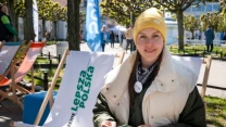 Młoda kobieta w żółtej czapce i jasnej kurtce siedzi na leżaku z napisem 'Lepsza Polska' w przestrzeni miejskiej, trzymając ulotkę, w tle widoczne są inne leżaki, flagi i przechodnie.