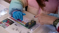 Dwie osoby wykonujące doświadczenie chemiczne, jedna z nich nosi niebieską rękawiczkę ochronną, na stole znajduje się zestaw probówek oraz karta z instrukcjami i kolorowymi okręgami.