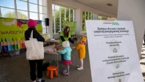 Grupa dzieci w kolorowych ubraniach uczestniczy w warsztatach pod dachem, rozmawia z osobą prowadzącą, w tle widoczny kolorowy baner i stanowisko z materiałami do zajęć, na pierwszym planie plansza informacyjna o warsztatach ekologicznych.