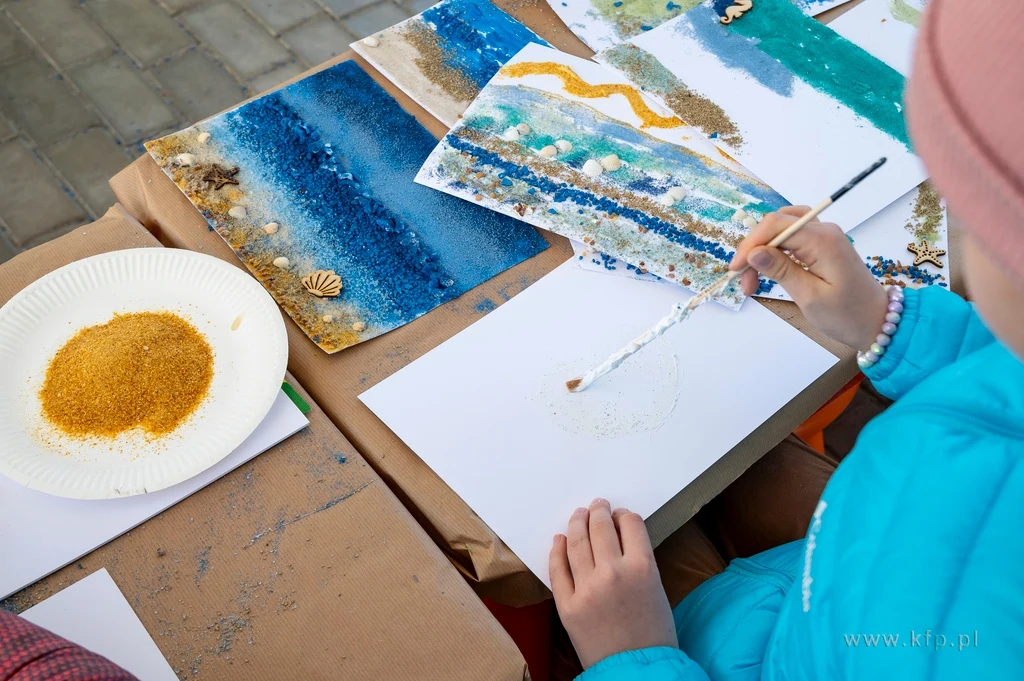 Czyste, Zielone Miasta w Sopocie fot. Anna Bobrowska/KFP/UMS Dziecko maluje obraz farbami na białej kartce, obok talerz z żółtym piaskiem oraz gotowe prace na stole, utrzymane w niebieskich i złotych barwach, przypominających morze.