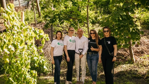 Grupa pięciu uśmiechniętych osób pozuje razem na tle młodych drzew i zieleni, wśród nich mężczyzna trzymający łopatę, sugerując udział w inicjatywie ekologicznej związanej z sadzeniem drzew.