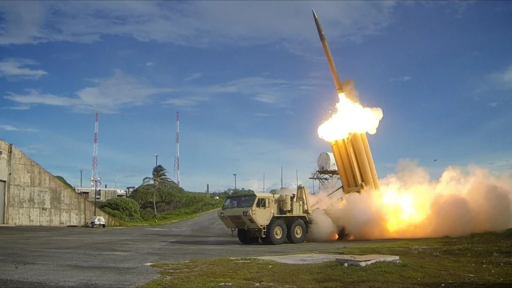 System THAAD podczas testów w 2013 roku Wojskowa wyrzutnia rakietowa odpalająca pocisk, unoszący się pionowo w powietrze. Obok betonowe umocnienia i stacja radarowa, całość w otwartym terenie z niebem pokrytym chmurami.