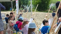 Grupa dzieci siedzi przy sztalugach na plaży i maluje obrazy pod opieką dorosłego, w tle drzewa i piasek, atmosfera spokojna i kreatywna.