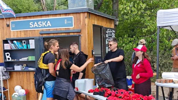 Grupa osób obsługiwana przy straganie z gadżetami nad wodą, na stołach maskotki, pojemniki i torby, w tle drewniany kiosk z napisem SASKA, otoczenie zielone, mające piknikowy charakter.